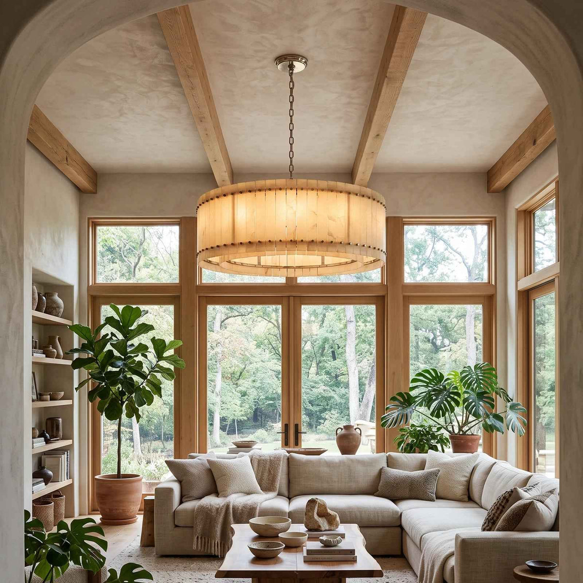 A large cylindrical drum pendant chandelier with a warm beige woven or fabric shade hangs in a bright, sunlit living room with wood beams, a sectional sofa, and potted plants.