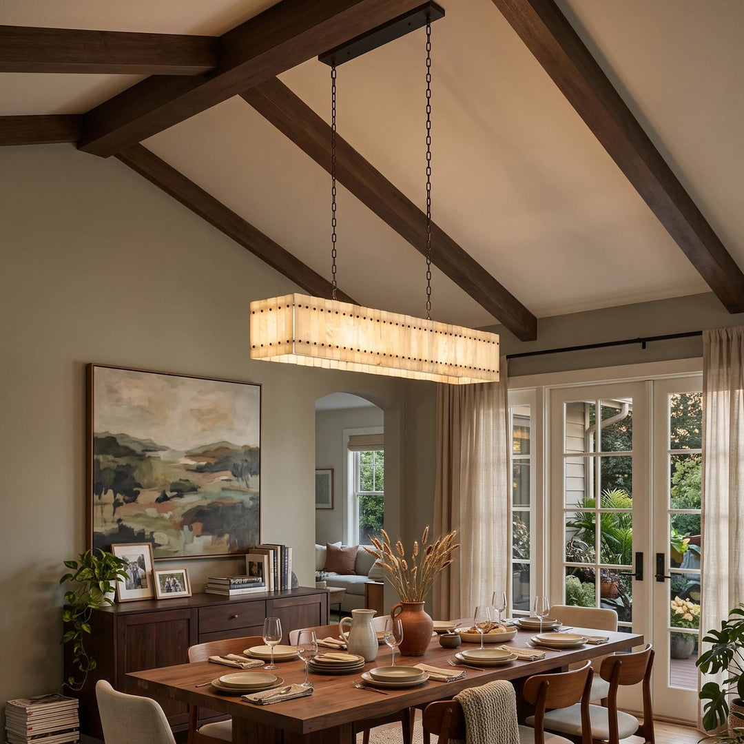 A linear chandelier with illuminated cream alabaster panels hangs above a wooden dining table beneath a vaulted ceiling with dark beams.