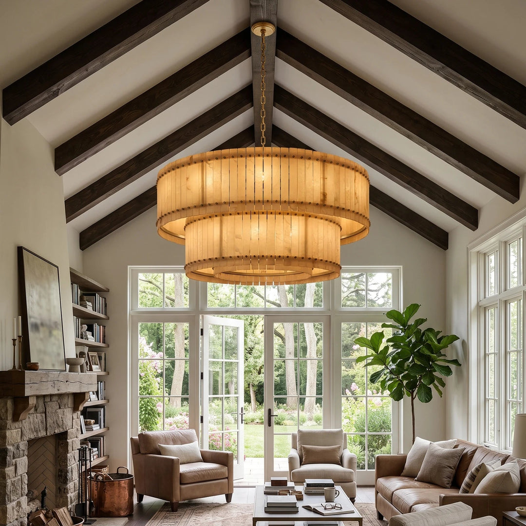 A large, tiered drum chandelier with a warm bamboo or wood slat shade hangs in a bright, high-ceilinged living room with exposed dark wood beams, large windows, and a stone fireplace.