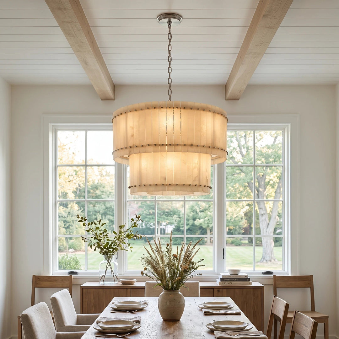 A two-tiered drum pendant chandelier with a warm beige fabric shade and brass-toned metal hardware, hanging in a bright, modern dining room with wood ceiling beams and large windows.
