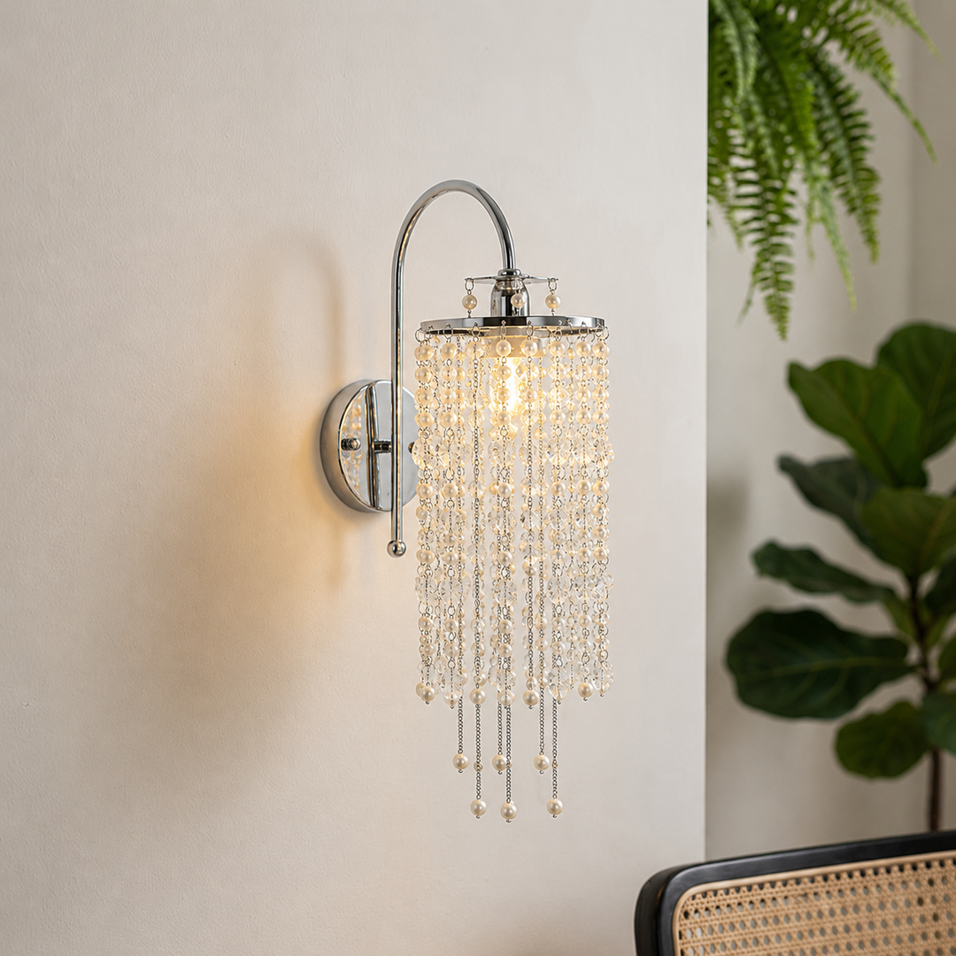 Polished chrome wall sconce with cascading crystal strands glows against a cream wall beside green plants and a cane-backed chair.