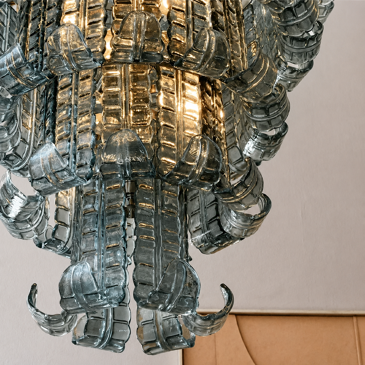 A close-up of a modern chandelier with textured, translucent smoky glass arms and a warm light glowing from within, set against a neutral beige wall.