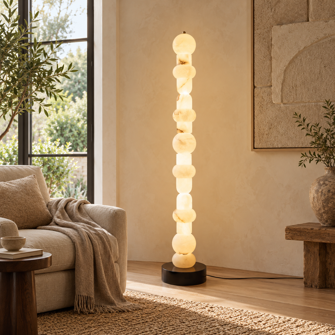 A modern floor lamp with a vertical stack of glowing oval bulbs on a black base, placed in a warm, neutral-toned living room next to a sofa and window.