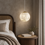 A spherical pendant light with a textured, translucent shell-like glass shade hangs over a bed in a styled bedroom. The light glows with a warm, soft light, while the room features a dark wood headboard, neutral bedding, a round side table with a book and small vase, and textured gray walls and curtains.