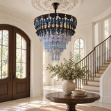 A large circular chandelier with blue and light blue hanging crystal-like beads and dark metal hardware, suspended in a bright entryway with a staircase, arched windows, and a round wooden table in the foreground.