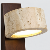 A modern cylindrical wall sconce with a textured beige travertine stone shade and a dark brown wooden mounting bracket, shown illuminated against a light gray background.