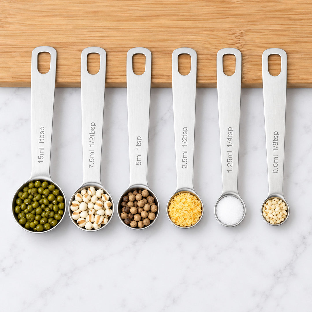 Six stainless-steel measuring spoons with grains and spices, shown fully from 0.6 to 15 ml.