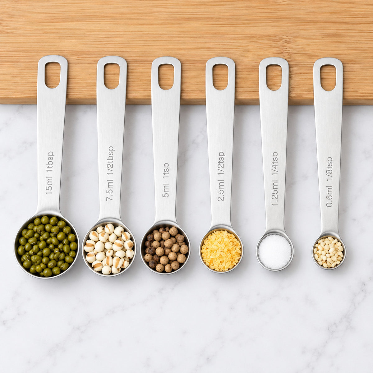 Six stainless-steel measuring spoons with grains and spices, shown fully from 0.6 to 15 ml.