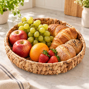 A round woven straw tray filled with fresh fruit and pastries, including apples, grapes, an orange, strawberries, a croissant, and a seeded bread, set on a light marble countertop in a bright kitchen.