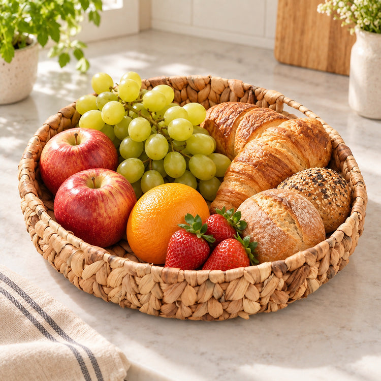 A round woven straw tray filled with fresh fruit and pastries, including apples, grapes, an orange, strawberries, a croissant, and a seeded bread, set on a light marble countertop in a bright kitchen.
