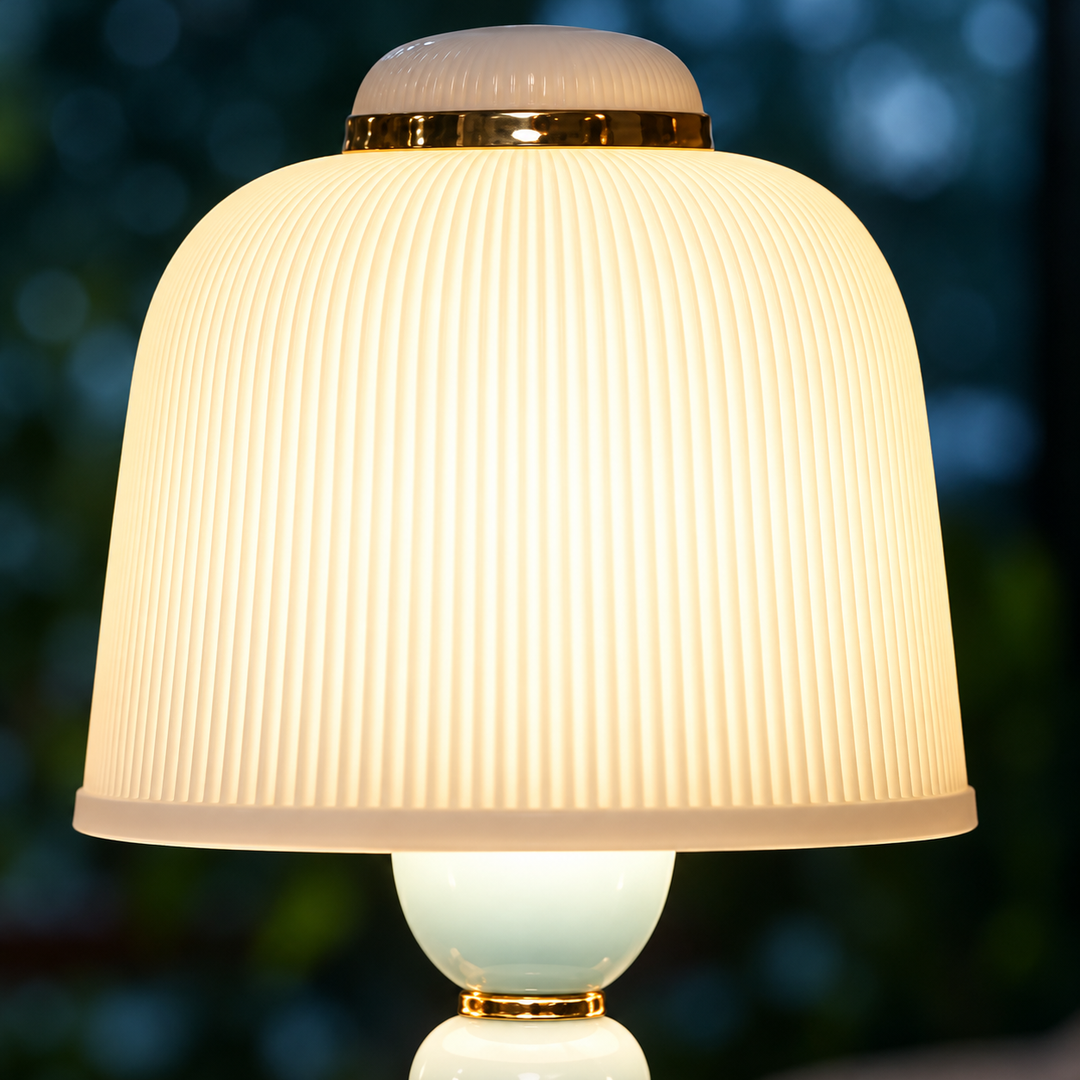 A table lamp with a ribbed white glass shade that glows warm yellow, a glossy white base, and gold metal trim, set against a blurred dark green background.
