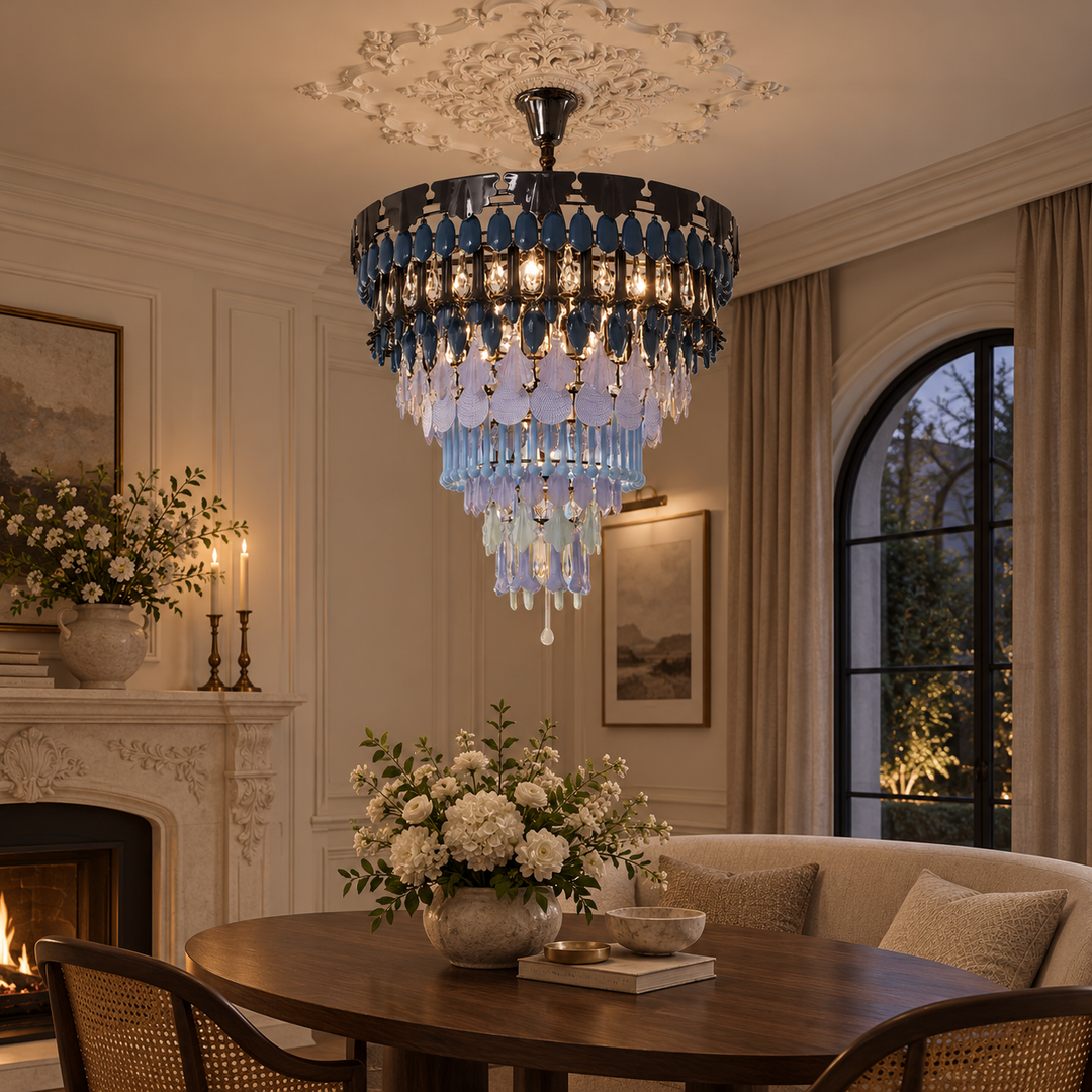 A luxury chandelier with cascading blue and clear crystal drops and a dark metal frame, hanging in an elegant, warmly lit dining room with a fireplace and wooden table.