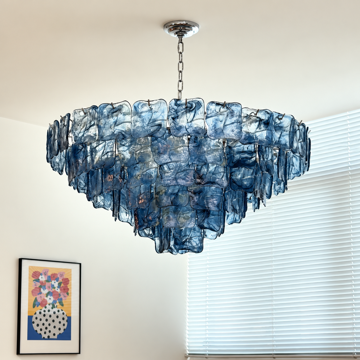A modern tiered chandelier made of textured, translucent blue glass panels suspended from a silver chain, shown hanging in a room with white blinds and a framed art print on the wall.