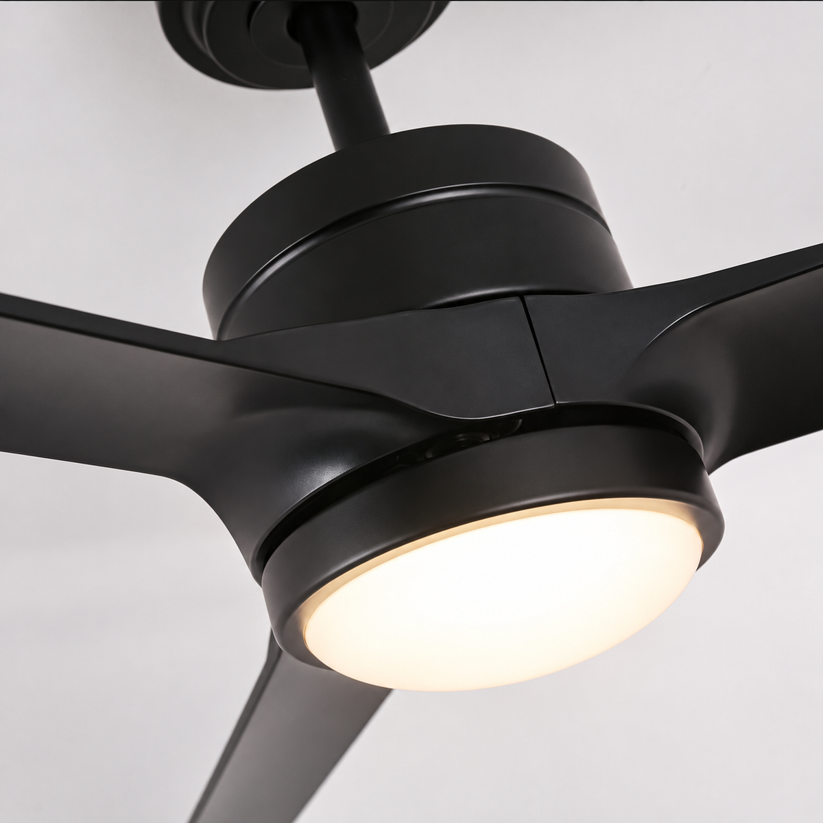 A close-up of a matte black modern ceiling fan with three blades and an integrated round light, shown against a plain light gray background.