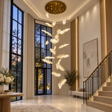 A modern multi-light cloud-shaped chandelier hanging in a luxurious high-ceilinged entryway with tall windows, a staircase, and warm ambient lighting.