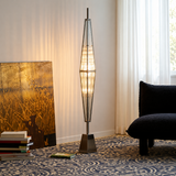 A modern geometric floor lamp with a warm glowing glass body and a dark metal frame, styled in a living room next to a dark armchair and a patterned blue rug.