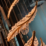 Large decorative hanging chandelier leaves in a warm gold-toned metal with clear textured glass, suspended in an indoor space with reflections and blue-toned walls.
