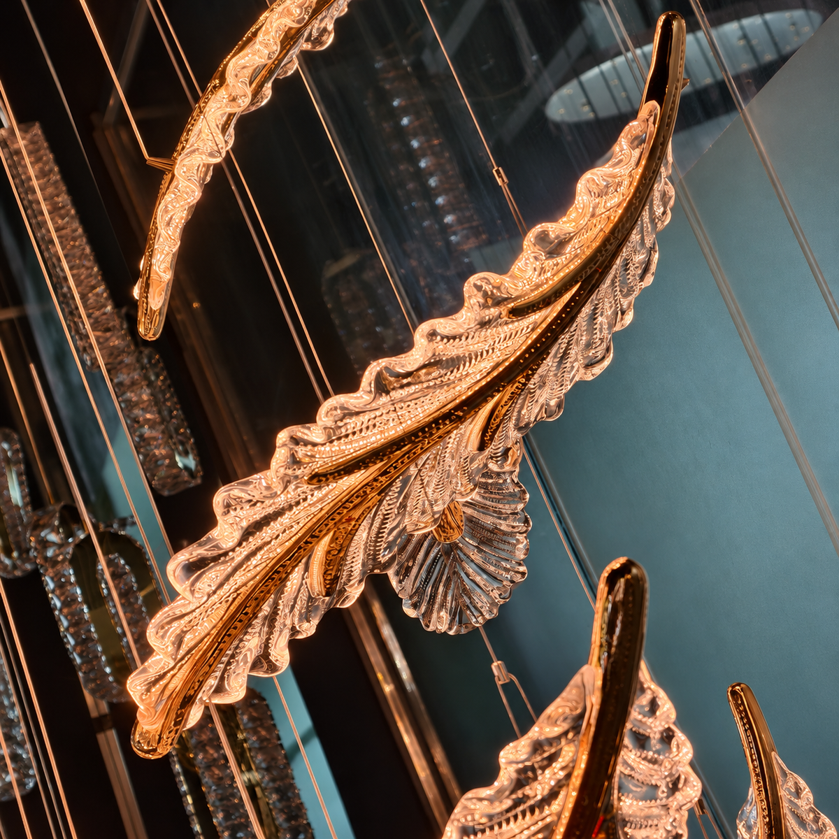 Large decorative hanging chandelier leaves in a warm gold-toned metal with clear textured glass, suspended in an indoor space with reflections and blue-toned walls.