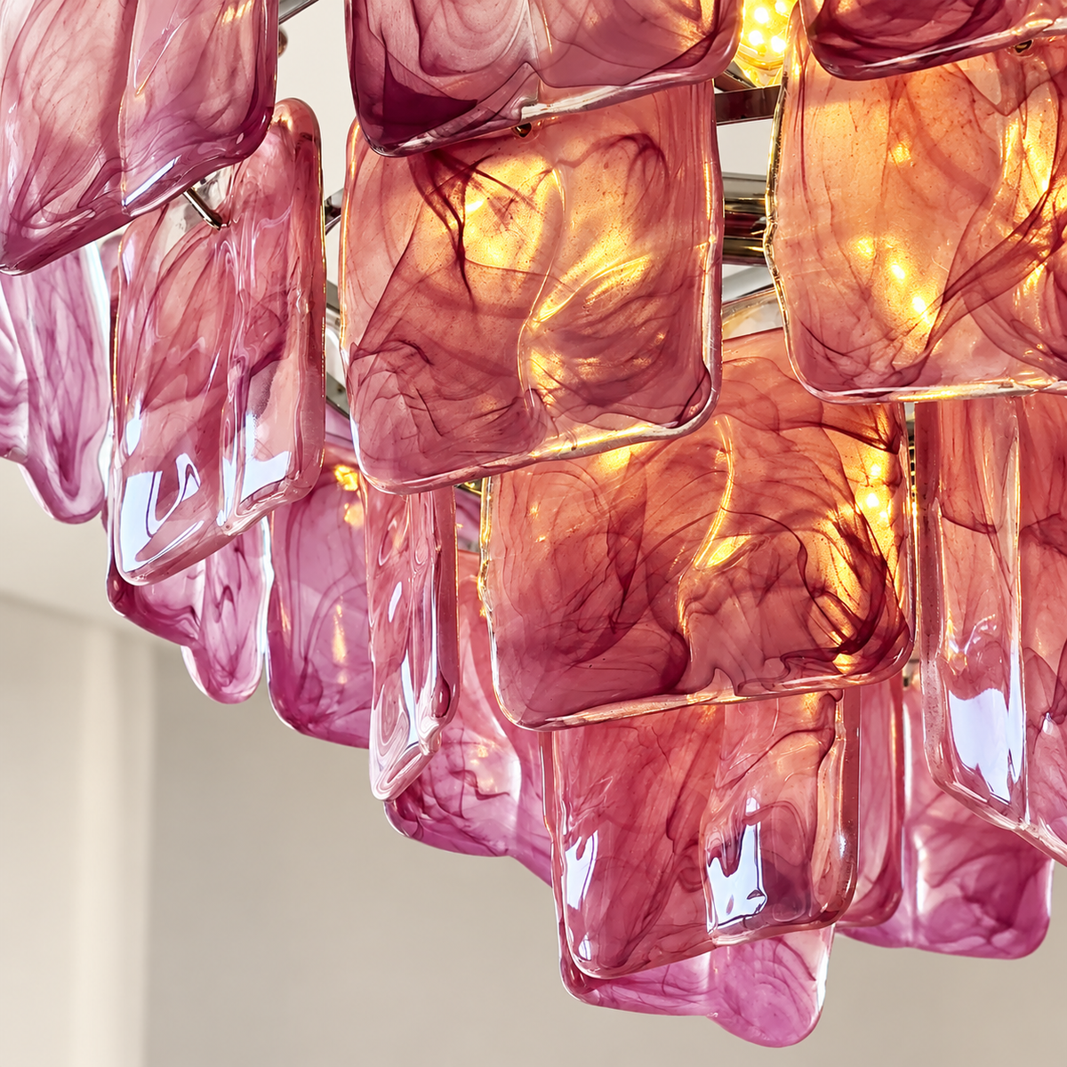 A close-up of a modern chandelier featuring multiple rectangular glass shades in a translucent pink and magenta color, illuminated from within to cast a warm golden glow.