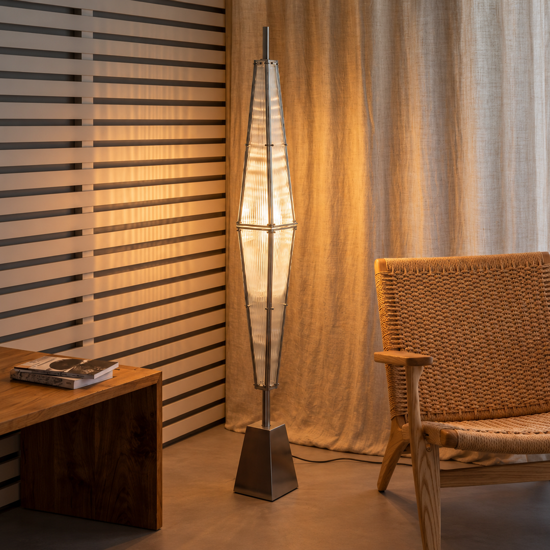 A modern floor lamp with a tall, tapered white ribbed shade and a dark metal base, standing in a warm-lit room next to a woven armchair and a slatted wooden wall panel.