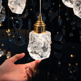 A hand supports one illuminated clear crystal shade of the chandelier, showing its irregular texture and polished gold cap against a dark background.