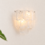 A modern wall sconce featuring a cluster of clear, textured glass blocks that glow warmly from an internal light source, mounted on a plain off-white wall.