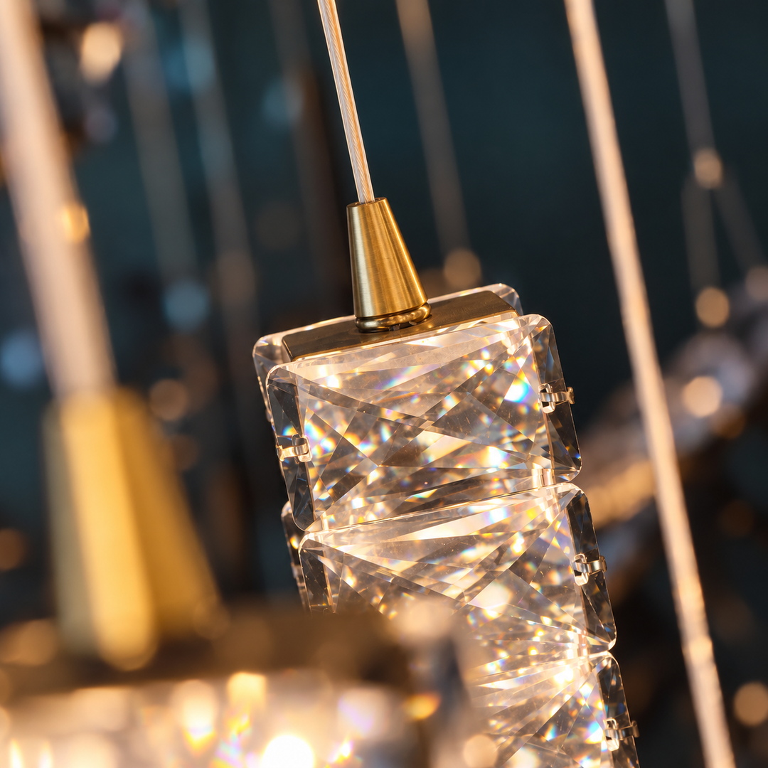 A close-up of a modern chandelier pendant with a faceted clear crystal or acrylic block and a brushed gold metal cap, suspended by a thin rod against a softly blurred dark background.
