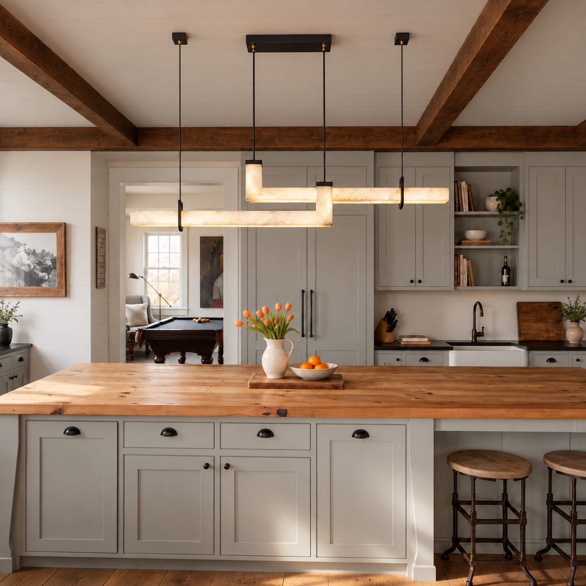 An illuminated alabaster linear pendant light hangs above a wooden kitchen island with gray cabinetry beneath exposed ceiling beams.