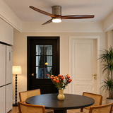 A modern ceiling fan with three dark wood blades and a cylindrical metal hub, mounted on a white ceiling in a dining room. The fan has an integrated light that glows downward.