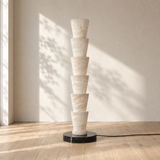 A modern stacked cone floor lamp made of stacked off-white marble cones on a round black base, set in a minimalist room with a light beige wall, wooden floor, and soft window shadows.