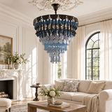 A large circular chandelier with cascading blue and clear glass teardrop crystals hangs in a bright, classic living room with a cream sofa, fireplace, and arched window.