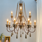 An ornate multi-arm chandelier with a distressed dark metal frame, wooden bead garlands, candle-style bulbs, and teardrop crystal drops, shown hanging against a light gray wall in a room setting.