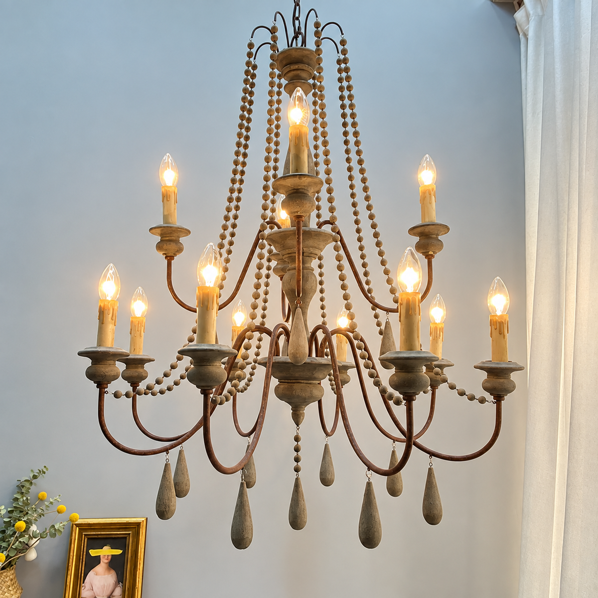 An ornate multi-arm chandelier with a distressed dark metal frame, wooden bead garlands, candle-style bulbs, and teardrop crystal drops, shown hanging against a light gray wall in a room setting.