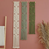 Three bohemian macrame wall hangings in cream, sage green, and olive green, hanging on a dusty pink wall with dried pampas grass and a wooden ladder prop.