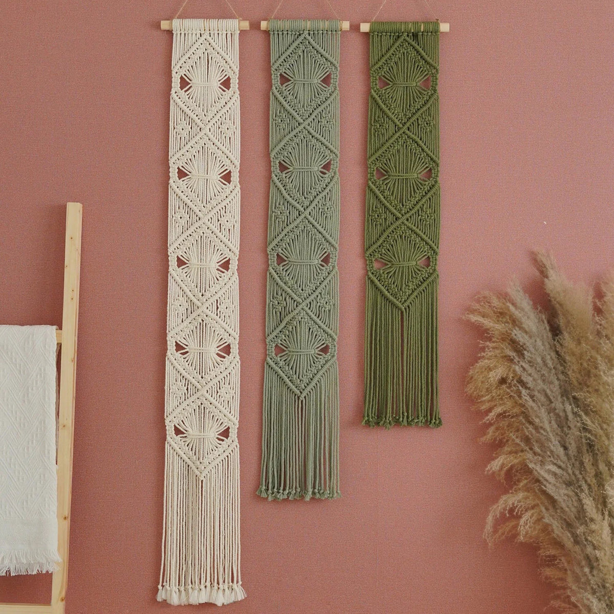 Three bohemian macrame wall hangings in cream, sage green, and olive green, hanging on a dusty pink wall with dried pampas grass and a wooden ladder prop.