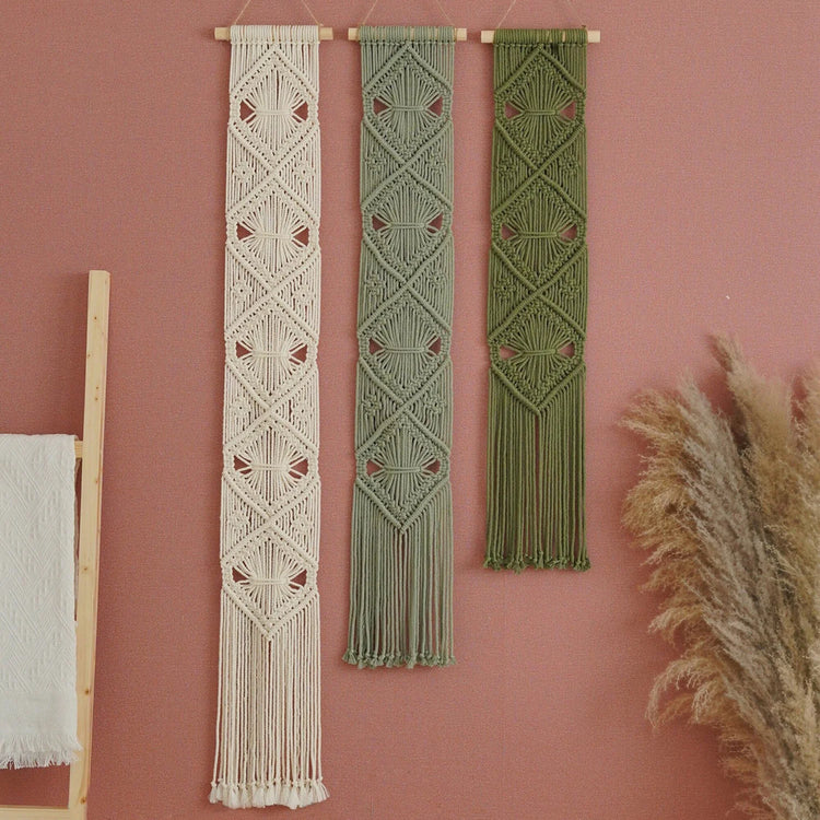 Three bohemian macrame wall hangings in cream, sage green, and olive green, hanging on a dusty pink wall with dried pampas grass and a wooden ladder prop.