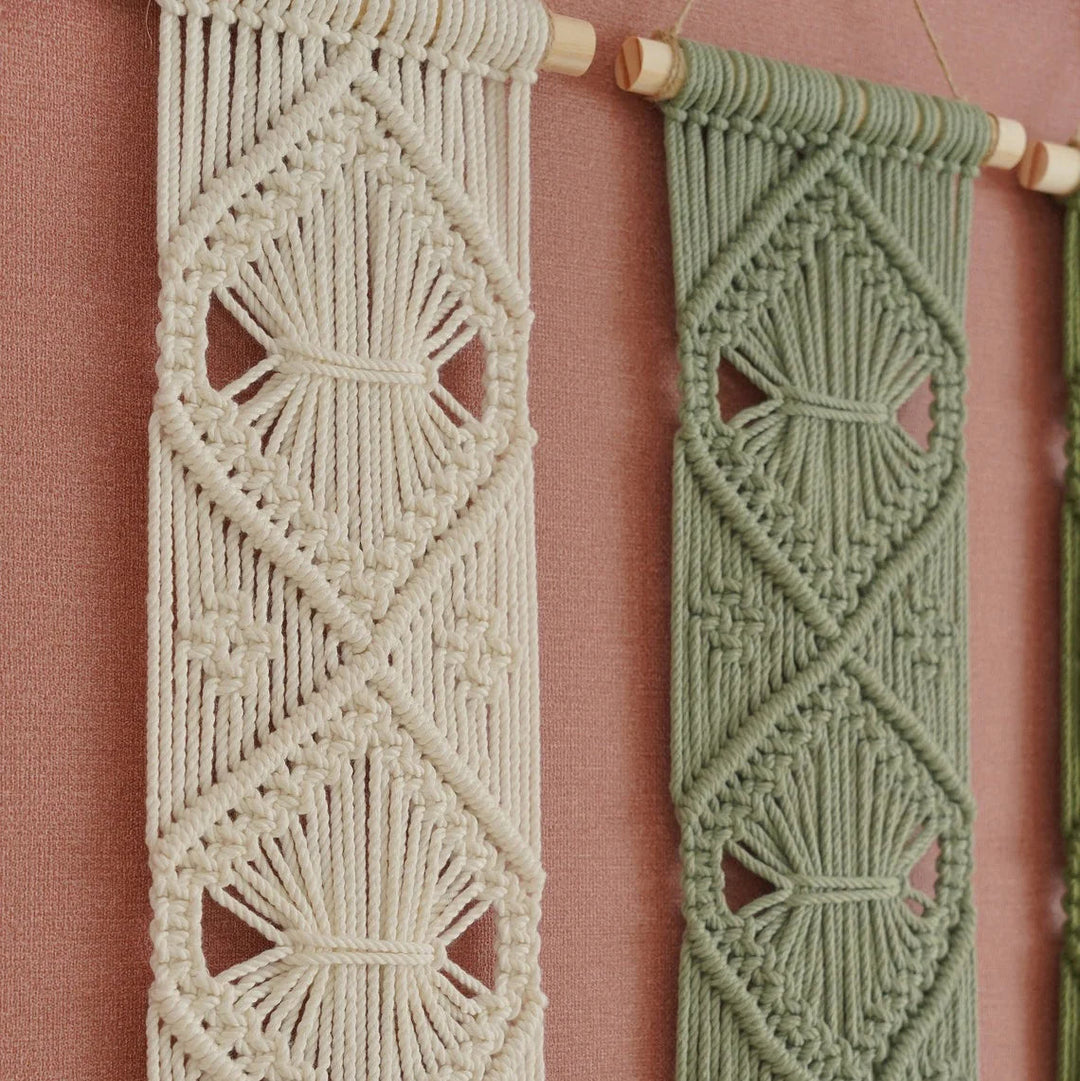 Two woven macrame wall hangings with geometric diamond patterns, one in cream and one in sage green, hanging from wooden dowels against a dusty pink wall.