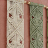 Two woven macrame wall hangings with geometric diamond patterns, one in cream and one in sage green, hanging from wooden dowels against a dusty pink wall.