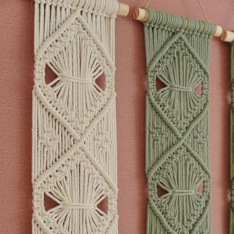 Two woven macrame wall hangings with geometric diamond patterns, one in cream and one in sage green, hanging from wooden dowels against a dusty pink wall.