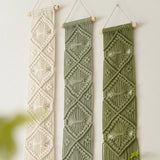 Three vertical macrame wall hangings hang from wooden dowels against a plain light wall. The left hanging is cream, the middle is sage green, and the right is olive green, each with geometric woven patterns and small cutout details.