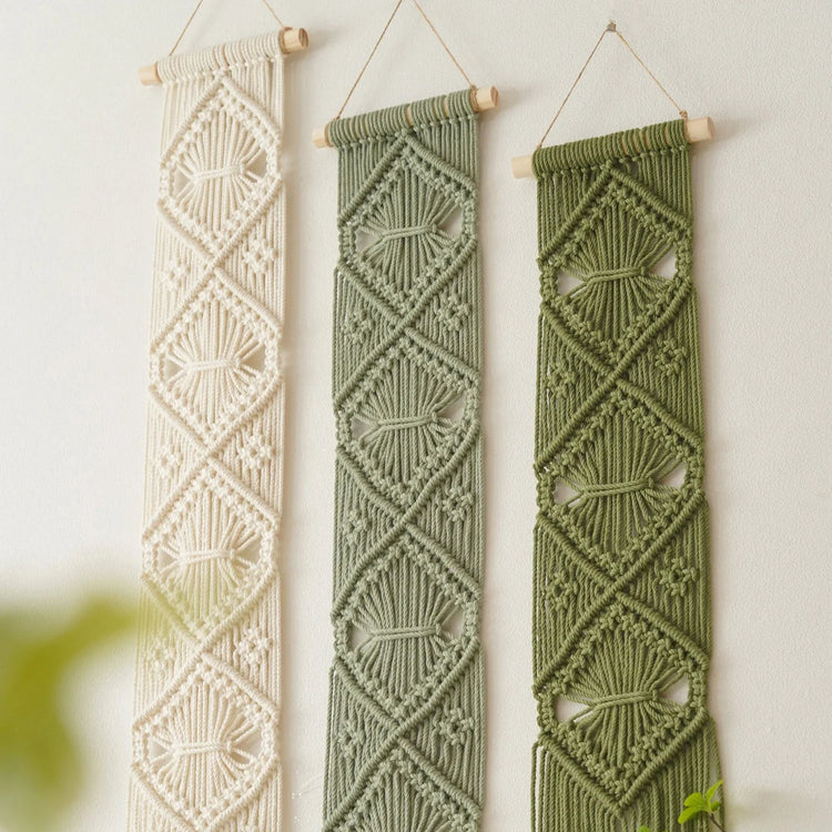 Three vertical macrame wall hangings hang from wooden dowels against a plain light wall. The left hanging is cream, the middle is sage green, and the right is olive green, each with geometric woven patterns and small cutout details.