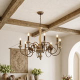 A rustic multi-light chandelier with a distressed metal frame and candle-style bulbs, hanging in a warm, traditional room with exposed wooden beams and a patterned wall hanging.