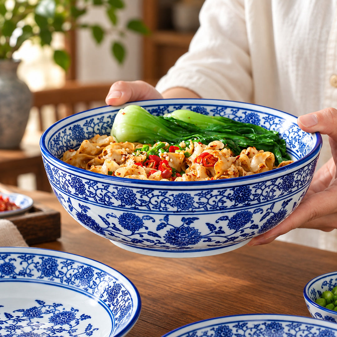 A person holds a large blue and white floral ceramic bowl filled with noodles, green vegetables, and red chili peppers, set on a wooden table with other matching bowls nearby.