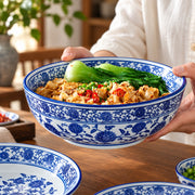 A person holds a large blue and white floral ceramic bowl filled with noodles, green vegetables, and red chili peppers, set on a wooden table with other matching bowls nearby.