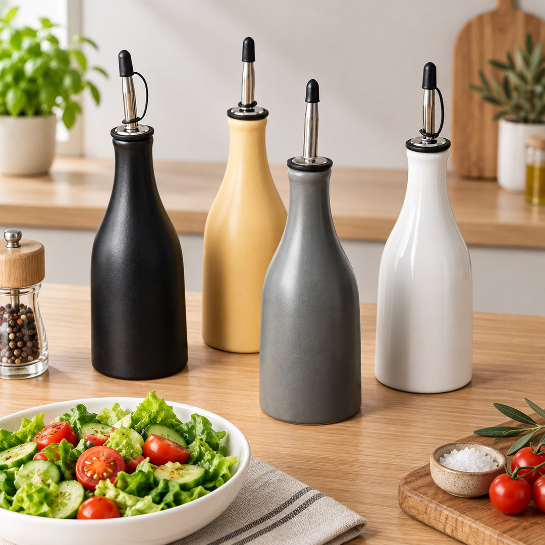 Four ceramic oil and vinegar bottles in matte black, mustard yellow, charcoal gray, and white, arranged on a wooden countertop with a salad bowl and kitchen decor in the background.