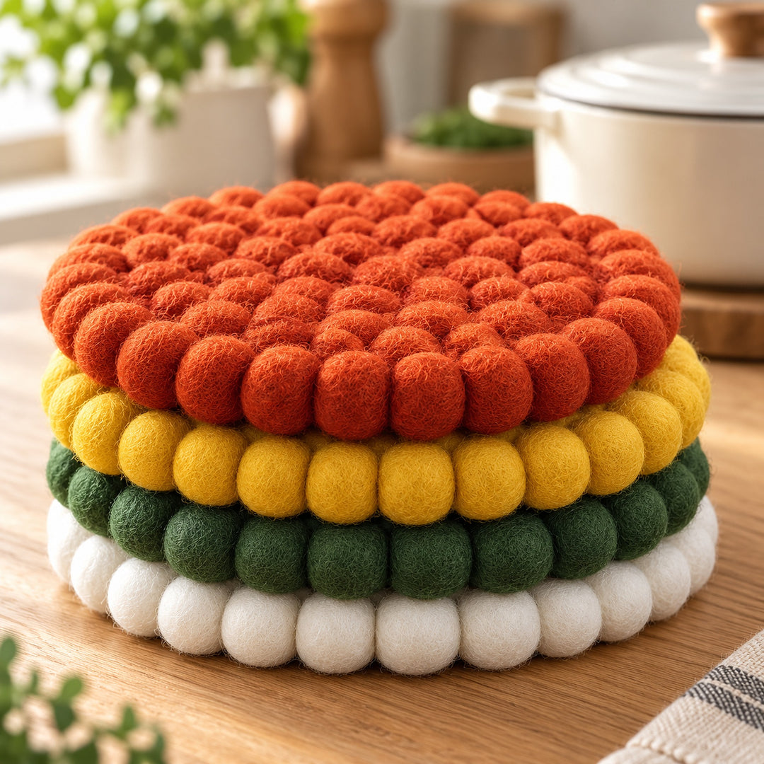 A round, multi-colored felt pom-pom coaster or trivet stacked on a wooden surface, featuring layers of white, green, yellow, and orange felt balls.