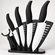 A 5-piece kitchen knife set with black handles and white blades, displayed in a black stand with a black peeler attached, on a light gray background.