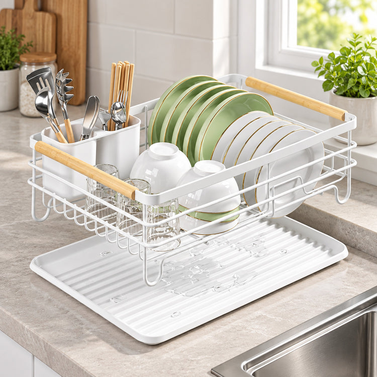 A white wire double-dish drier rack with light wood handles and a white drain tray, shown on a kitchen counter with plates, bowls, and utensils. The background features a tiled wall, a window, and potted plants.
