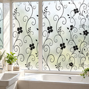 Black floral scroll vine window film applied to frosted glass panels in a bright bathroom, with a bathtub, sink, and potted plants visible.