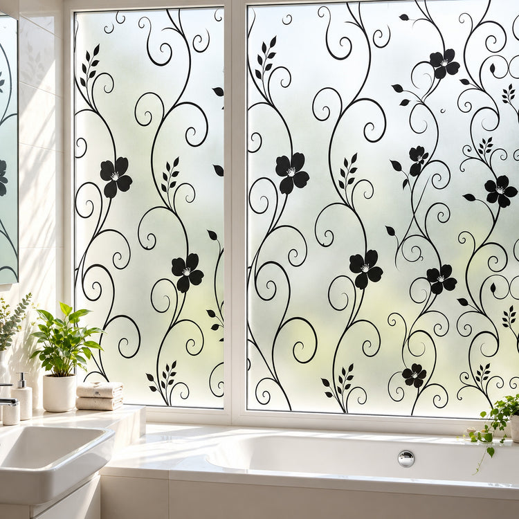Black floral scroll vine window film applied to frosted glass panels in a bright bathroom, with a bathtub, sink, and potted plants visible.